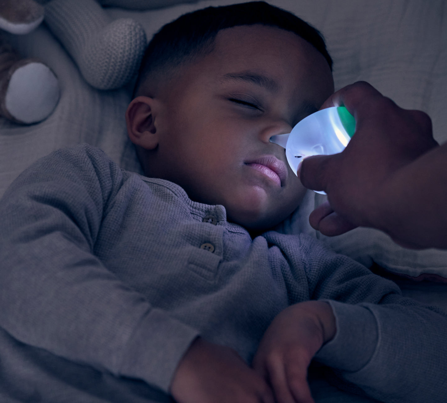 Child lying down with a hand holding a light up nasal bulb close to their face in a dimly lit room.